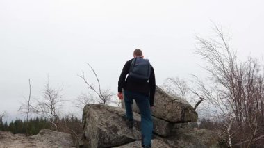 Sırt çantalı genç adam kayanın üstünde yürüyor ve cep telefonuyla selfie çekiyor.