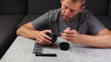 Masanın arkasında oturan ve tamir eden bir adam kırık kamera lenslerini tornavidayla temizliyor.