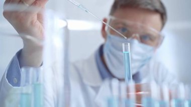 Güvenlik gözlüklü ve mavi maskeli bir adam laboratuvar tüpleriyle çalışıyor. Modern bir laboratuarda, yakın plan bir araştırmacıyı kullanıyor. Bilim ve tıp.