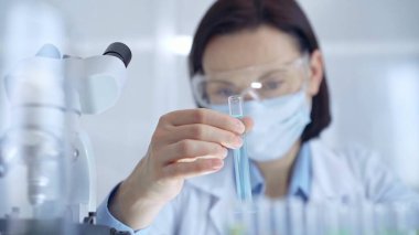 Koruyucu gözlükleri ve maskesiyle laboratuvar tüpünü mavi sıvıyla inceleyen bilim adamı kadın araştırmacı. Mikrobiyoloji ve bilim konsepti.