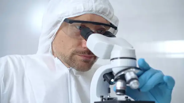 stock image Scientist in ppe analyzing samples with microscope. Close-up of a focused researcher in a lab coat and protective gear using a microscope in a laboratory setting.