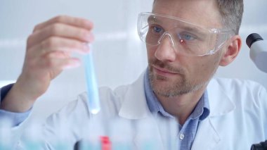 Koruyucu gözlüklü bilim adamı mavi bir sıvıyla laboratuvar tüpünü inceliyor. Mikrobiyoloji ve bilim konsepti.