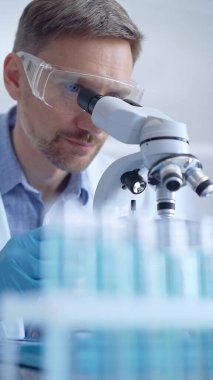 Koruyucu gözlüklü ve eldivenli bilim adamı laboratuarda mikroskop kullanıyor, araştırmacının dikey portresi. Mikrobiyoloji bilim konsepti.