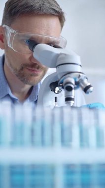Koruyucu gözlüklü ve eldivenli bilim adamı laboratuarda mikroskop kullanıyor, araştırmacının dikey portresi. Mikrobiyoloji bilim konsepti.