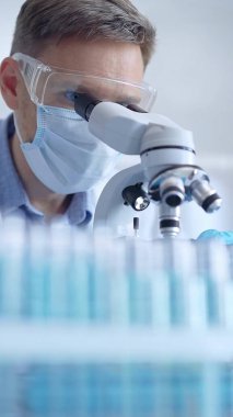 Koruyucu gözlüklü, maskeli ve eldivenli bilim adamı laboratuarda mikroskop kullanıyor, araştırmacının dikey portresi. Mikrobiyoloji ve bilim.