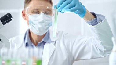 Koruyucu eldivenleri ve gözlükleri olan bir bilim adamı yeşil sıvıyla laboratuvar tüpünü inceliyor. Mikrobiyoloji ve bilim konsepti.