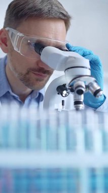 Koruyucu gözlüklü ve eldivenli bilim adamı laboratuarda mikroskop kullanıyor, araştırmacının dikey portresi. Mikrobiyoloji bilim konsepti.