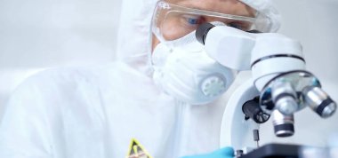 Koruyucu ekipman kullanan profesyonel bilim adamı bir laboratuvar ortamında mikroskop kullanıyor, yakın plan. Bilim ve tıp kavramları.