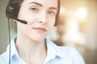 Kulaklığı olan güzel bir kadın uzman müşterilere çevrimiçi yardım ediyor ve güneşli çağrı merkezinde büyüleyici bir şekilde gülümsüyor. İş adamları konsepti.