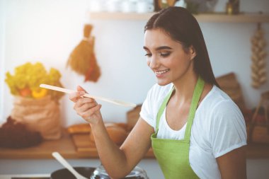 Çekici, genç, siyah saçlı bir kadın, güneşli mutfakta ayakta ve gülümserken tahta kaşıkla sıcak yemek tadıyor. Yemek pişirme ve ev idaresi kavramları.