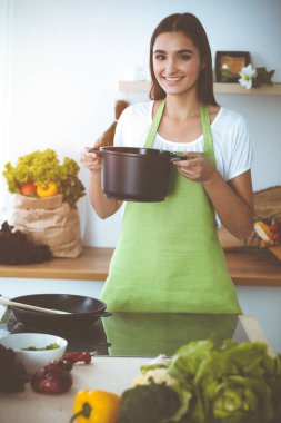 Çekici, genç, koyu saçlı bir kadın, güneşli mutfakta ayakta durup gülümserken yeni keto tarifiyle çorba hazırlıyor. Yemek pişirme ve ev idaresi kavramları.