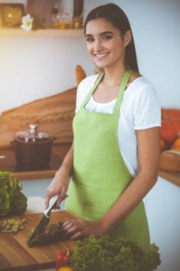 Çekici, genç, koyu saçlı bir kadın güneşli mutfakta ayakta durup gülümserken lezzetli taze vejetaryen salatası hazırlıyor. Yemek pişirme ve ev idaresi kavramları.