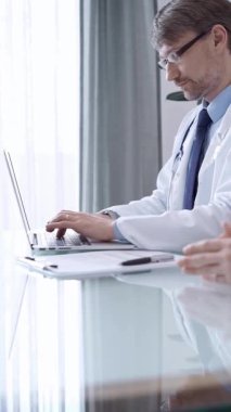 Beyaz önlüklü erkek doktor laptoptaki hasta bilgilerini incelerken modern bir klinikteki laptop verilerine odaklandı. Tıp konsepti.