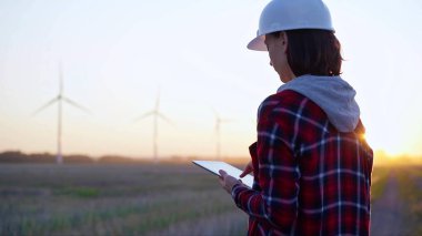Kadın mühendis, güneş batarken rüzgar türbinleriyle dolu bir alanda tablet bilgisayarla not alıyor. Temiz enerji ve mühendislik kavramı.