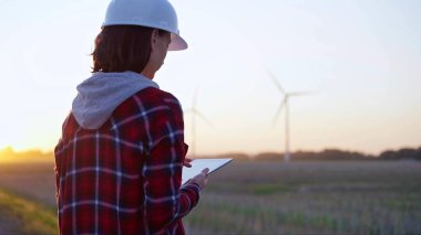 Kadın mühendis, güneş batarken rüzgar türbinleriyle dolu bir alanda tablet bilgisayarla not alıyor. Temiz enerji ve mühendislik denetimi kavramı.