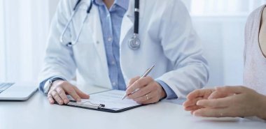 Bir doktor ve bir hasta. Mavi bir gömleğin üzerine beyaz bir ceket giyen hekim klinikteki muayene sırasında tıbbi kayıt formu dolduruyor. Tıbbi hizmet.