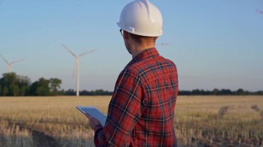 İş yerindeki mühendis beyaz bir koruyucu kask takıyor ve güneş batarken rüzgar türbinli bir alanda tablet bilgisayarla notlar alıyor. Temiz enerji kavramı.