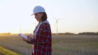 Beyaz fıçı giyen yetişkin kadın mühendis güneş batarken rüzgar türbinleriyle dolu bir alanda bir panoya notlar alıyor..