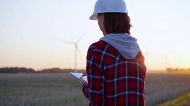 Kadın mühendis, güneş batarken rüzgar türbinleriyle dolu bir alanda tablet bilgisayarla not alıyor. Temiz enerji ve mühendislik kavramı.