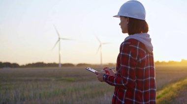 Beyaz fıçı takan yetişkin kadın mühendis rüzgar türbinleriyle dolu bir alanda not alıyor..