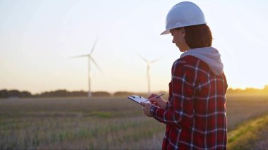 Beyaz fıçı giyen yetişkin kadın mühendis rüzgar türbinleriyle dolu bir alanda not alıyor. Güneş batarken, arka görüş....