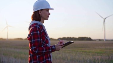 Beyaz fıçı takan yetişkin kadın mühendis rüzgar türbinleriyle dolu bir alanda not alıyor..