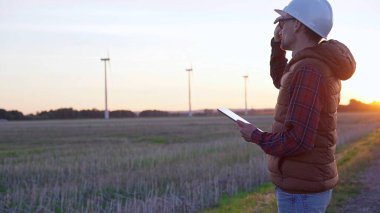 Beyaz koruyucu kask takan mühendis güneş batarken rüzgar türbinli bir alanda tablet bilgisayarla not alıyor. Temiz enerji ve mühendislik kavramı.