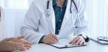 Doktor ve bir hasta. Yeşil gömleğin üzerine beyaz bir tıbbi önlük giyen hekim klinikteki muayene sırasında tıbbi kayıt formu dolduruyor. Tıbbi hizmet.