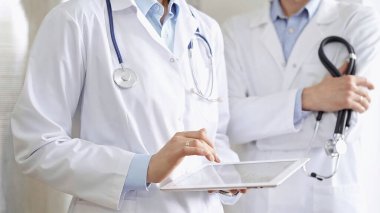 Dijital tablet kullanan, beyaz önlük ve steteskop giyen iki doktor bir hastanın tıbbi geçmişini tartışıyor. Tıp ve Bilim.