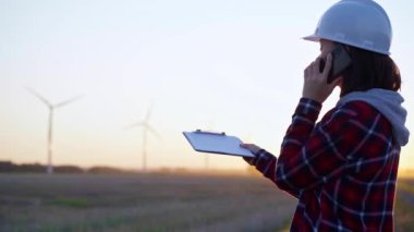 Kadın mühendis, güneş batarken rüzgâr türbinleriyle telefonda konuşuyor. Temiz enerji ve mühendislik denetimi kavramı.