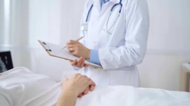 Kadın doktor hastane yatağında yatan bir hastanın başında dikilirken not alıyor.