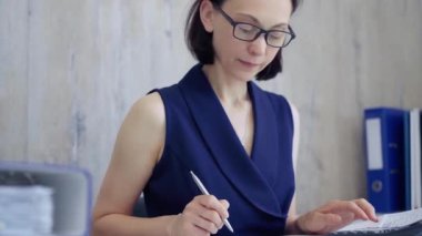 İş kadını ofisteki masasında kağıt işleriyle uğraşıyor, hesap makinesi kullanıyor ve not alıyor. Vergiler, iş denetimleri.