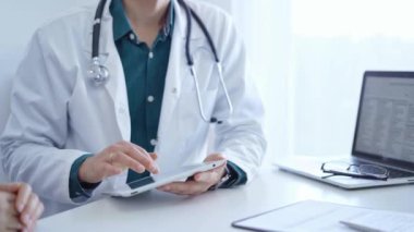 Doktor ve bir hasta. Yeşil gömleğin üzerine beyaz bir tıbbi önlük giyen hekim klinikteki muayene sırasında tablet bilgisayar kullanıyor. Tıp konsepti.