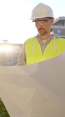 Erkek ve kadın mimarlar veya mühendisler güvenlik kasketleri ve yelekleri giyip şemaları tutuyorlar ve gün doğumunda inşaat alanında bir şey tartışıyorlar, ön dikey görüş. Mimarlık konsepti.