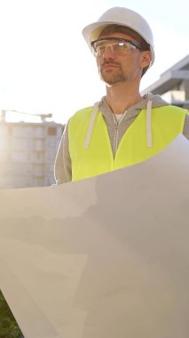 Erkek ve kadın mimarlar veya mühendisler güvenlik kasketleri ve yelekleri giyip şemaları tutuyorlar ve gün doğumunda inşaat alanında bir şey tartışıyorlar, ön dikey görüş. Mimarlık konsepti.