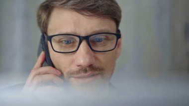 Gözlüklü bir iş adamı telefonda konuşuyor. Bulanık dizüstü bilgisayarla kısmen gizlenmiş. Ön görünüm ve yakın çerçeve.