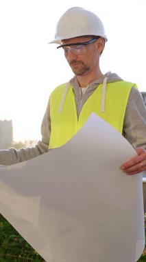 Erkek ve kadın mimarlar veya mühendisler güvenlik kasketleri ve yelekleri giyip şemaları tutuyorlar ve gün doğumunda inşaat alanında bir şey tartışıyorlar, ön dikey görüş. Mimarlık konsepti.