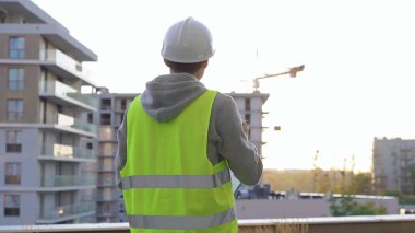 Beyaz koruyucu miğferi ve güvenlik yeleği olan erkek inşaat mühendisi gün batımında bir inşaat alanını incelerken panoya yazı yazıyor. Mimar konsepti.