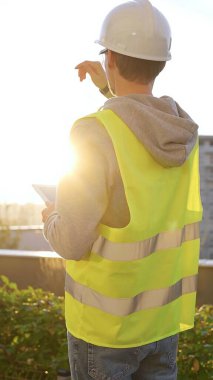 Beyaz miğfer ve güvenlik yeleği olan bir mimar ya da mühendis, gün doğumunda dikey olarak bir inşaat alanını incelerken dijital tablete notlar alıyor. Mimarlık konsepti.