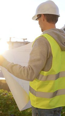 Güvenlik kasketi ve yeleği olan bir mimar ya da mühendis elinde planla gün doğumunda dikey olarak inşaat alanı arıyor. Mimarlık ve mühendislik.