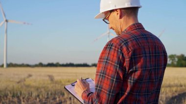 İş yerindeki mühendis beyaz bir koruyucu kask takıyor ve güneş batarken rüzgar türbinleriyle dolu bir alanda not alıyor. Temiz enerji kavramı.