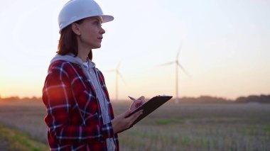 Kadın mühendis, güneş batarken rüzgâr türbinleriyle dolu bir alanda bir panoya not alıyor. Temiz enerji ve mühendislik denetimi kavramı.