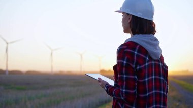 Kadın mühendis, güneş batarken rüzgar türbinleriyle dolu bir alanda tablet bilgisayarla not alıyor. Temiz enerji ve mühendislik kavramı.