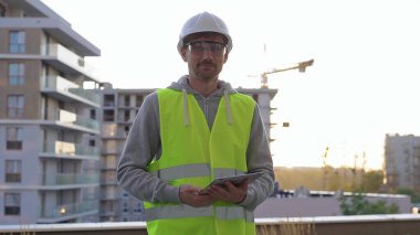 İnşaat şantiyesi mühendisi elinde tablet tutuyor ve sabah erken saatlerde arka planda güneş doğarken sıkı şapka ve güvenlik yeleği giyiyor..
