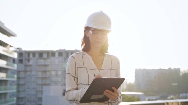 Beyaz işaretli bluz ve kasketli şapka giyen kadın inşaat mühendisi, gün batımında bir inşaat alanını incelerken not alıyor. Mimarlık ve mühendislik kavramları.