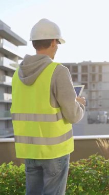 Beyaz miğfer ve güvenlik yeleği takan mühendis, gün doğumunda bir inşaat alanını dikey olarak incelerken tablet bilgisayar kullanıyor..