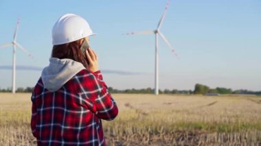 Beyaz koruyucu kask takan kadın mühendis güneş batarken rüzgar türbinleriyle dolu bir alanda akıllı bir telefonla konuşuyor. Temiz enerji ve mühendislik denetimi kavramı.