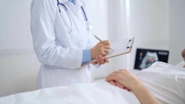 Kadın doktor hastane yatağında yatan bir hastanın başında dikilirken not alıyor.
