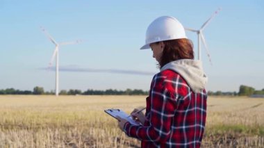 Beyaz koruyucu kask takan kadın mühendis güneş batarken rüzgâr türbinli bir alanda not alıyor. Temiz enerji ve mühendislik kavramı.
