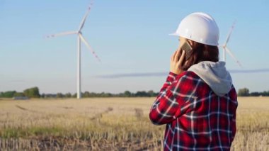 Beyaz koruyucu kask takan kadın mühendis güneş batarken rüzgar türbinleriyle dolu bir alanda akıllı bir telefonla konuşuyor. Temiz enerji ve mühendislik denetimi kavramı.